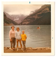 three women standing next to each other in front of a body of water with mountains in the background