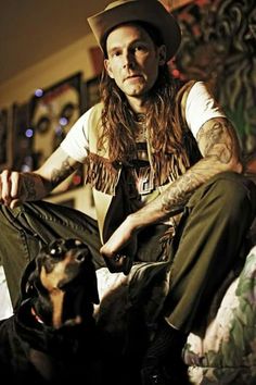 a man with long hair sitting on a couch next to a black and brown dog