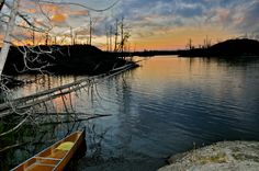 a canoe sitting on the shore of a lake at sunset with trees in the foreground