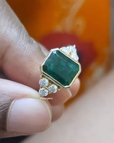 a close up of a person's hand holding an emerald and diamond ring