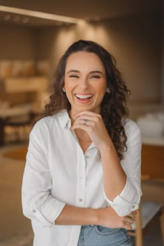 a smiling woman in a white shirt and jeans stands with her hands on her chin