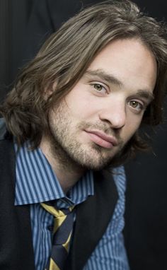 a man with long hair wearing a vest and striped shirt is looking at the camera