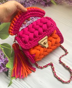 a hand holding a pink and orange purse with tassels on the handle, next to purple flowers