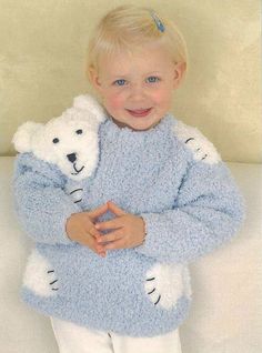 a little boy holding a teddy bear in his arms while wearing a blue knitted sweater