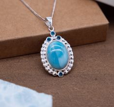 Discover the epitome of sophistication with our Oval Silver Pendant featuring a captivating Larimar center stone. This pendant seamlessly marries classic design with intricate detailing, creating an accessory that transcends trends. Oval Larimar Centerpiece: The focal point of this pendant is an exquisite oval Larimar stone, celebrated for its serene blue hues. Embraced by a silver setting, the Larimar takes center stage, infusing the piece with a tranquil and timeless allure. Intricate Heart-Shaped Frame: Surrounding the Larimar is a meticulously crafted oval silver frame. Comprising an array of small silver heart shapes interconnected in an artistic pattern, this frame adds a touch of romance and femininity to the design. Blue Topaz Accents: Three blue topaz stones are delicately studded Elegant Larimar Pendant Jewelry, Blue Topaz Oval Pendant Necklace For Anniversary, Oval Blue Topaz Birthstone Necklace, Elegant Larimar Cabochon Jewelry, Elegant Round Larimar Jewelry, Elegant Larimar Jewelry For Formal Events, Blue Topaz Necklaces With Accent Stones For Anniversary, Blue Topaz Gemstones For Gifts, Anniversary Blue Topaz Necklaces With Accent Stones