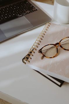 a pair of glasses sitting on top of a notebook next to a cup and laptop computer