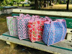 four bags are lined up on a bench in the park, one is pink and two are blue