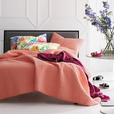 a bed covered in an orange blanket next to a vase with purple flowers on it