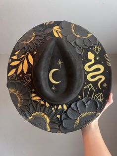 Halloween hat Brim is about 3.75" wide  Content: Vegan Suede Adjustable to fit your head better if needed  Elevate your style with this stunning hand-painted wide-brim fedora, crafted from luxurious vegan suede in a timeless black and gold palette. This one-of-a-kind piece is adorned with intricate designs featuring a serpent winding gracefully around the crown, sparkling crystals, celestial stars, delicate leaves, and a crescent moon. Each flower is meticulously painted with thick, textured strokes, adding a unique tactile dimension to the hat's sophisticated design. This fedora isn't just a hat; it's a wearable work of art. Perfect for making a statement at festivals, special events, or just adding a touch of magic to your everyday outfits. Whether you're dressing up or down, this versat Black Curved Brim Fedora For Halloween, Custom Curved Brim Costume Hat For Festivals, Custom Curved Brim Festival Costume Hat, Custom Costume Hat With Curved Brim For Festivals, Custom Festival Costume Hat With Curved Brim, Adjustable Fedora Felt Hat For Halloween, Adjustable Flat Brim Halloween Costume Hat, Black Brimmed Fedora For Halloween, Adjustable Flat Brim Hat For Halloween