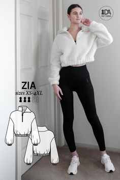 a woman standing in front of a door wearing black pants and a white sweater with zippers