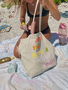 a woman is sitting on the beach with a tote bag in her hand and some other items around her