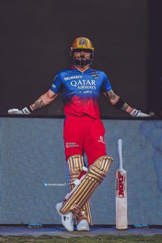 a man in red and blue uniform standing next to a cricket bat with his arms outstretched