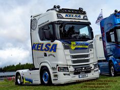 two semi trucks parked next to each other on a grass covered field with trees in the background