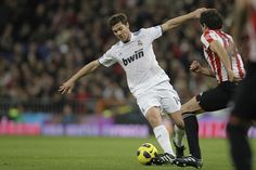 two men playing soccer on a field with people watching from the stands in the background