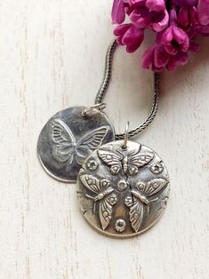 two necklaces with butterflies on them sitting next to purple flowers and one is silver