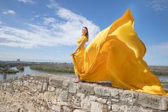 a woman in a yellow dress standing on a stone wall next to a body of water