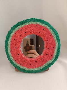 a hand holding a cell phone in front of a watermelon shaped mirror on a white surface