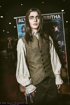 a man with long hair wearing a vest and tie standing in front of a poster