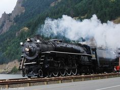 an old fashioned steam train is traveling down the tracks near a river and some mountains