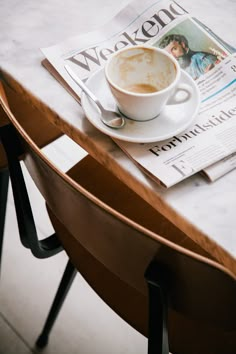 a cup of coffee sitting on top of a wooden table next to a newspaper and chair