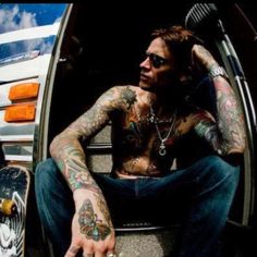 a man with tattoos sitting in the back of a truck next to a skateboard