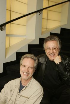 two men sitting next to each other in front of a stair case, smiling at the camera