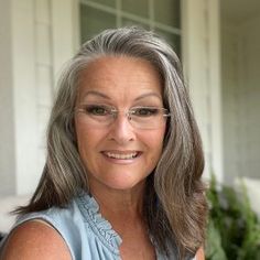 an older woman with gray hair and glasses smiling for the camera in front of a house