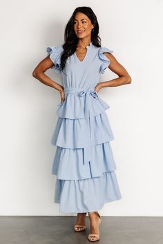 a woman wearing a blue dress with ruffles on the skirt and heels standing in front of a white wall