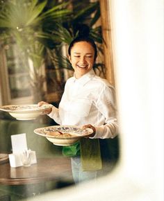 a woman holding two plates with food on them in front of a window and smiling at the camera