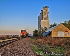 a train traveling down tracks next to a tall building and grass covered field with trees in the foreground