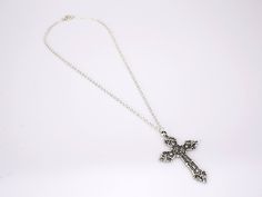 The cross pendant of this lovely piece is an elegantly crafted piece with many charming accents adorning it. It's forged from a strong 316L stainless steel material and finished in a polished silver coat. The exterior border and detailed outlines on this piece are smooth and clean-cut, while the interior India is covered in a rougher, granite-like surface. The contrasting textures are clear and highlight the details on the piece beautifully. The cross stands at 56 mm long. This Innovato Design piece comes with a 18" silver twisted Singapore chain.  Product Highlights:    Made from durable stainless steel silver  Elegant Cross Pendant Design  Twisted Singapore Chain Necklace (Length: 18") Metal Cross Pendant Necklace, Silver Cross Pendant Jewelry, Elegant Metal Crucifix Cross Necklace, Spiritual Metal Cross Necklace, Silver Chain Cross Jewelry, Silver Chain Metal Cross Jewelry, Silver Chain Cross Metal Jewelry, Metal Cross Jewelry With Silver Chain, White Gold Metal Cross Pendant Jewelry