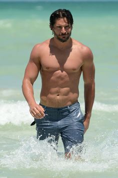 a shirtless man standing on the beach with his surfboard