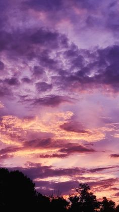 the sky is filled with purple clouds as the sun sets over some trees in the distance