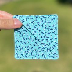 a hand holding up a small square piece of fabric with blue flowers on it and a green field in the background