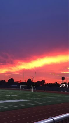 the sun is setting over a soccer field with trees in the background and purple clouds