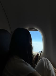 a woman sitting in an airplane looking out the window at the sky and reading a book