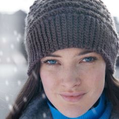 a close up of a person wearing a knitted hat and blue scarf with snow falling on the ground