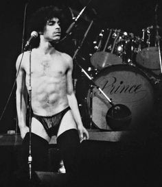 a shirtless man standing in front of a microphone on stage with drums behind him