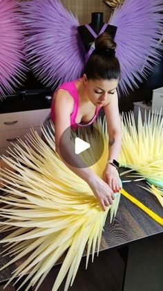 a woman in pink shirt working on large yellow and purple feathered object with scissors