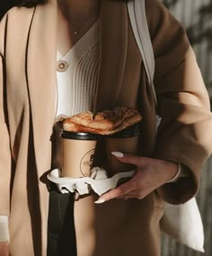 a woman holding a coffee cup and some food in her hand while wearing a brown coat