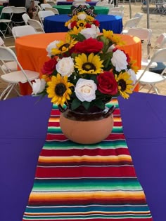 a flower pot with sunflowers and roses in it on a colorful table cloth