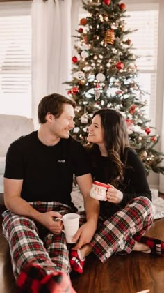 a man and woman are sitting on the floor in front of a christmas tree holding coffee cups