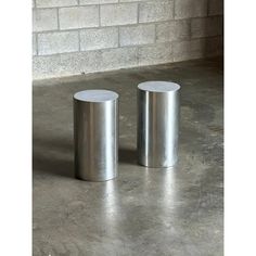 two stainless steel stools sitting next to each other on a cement floor in front of a brick wall