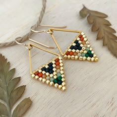 the triangle earrings are made with gold and multi - colored seed beaded beads, on a white wooden surface