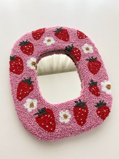 a pink knitted mirror with strawberries and daisies on the front, sitting on a white surface