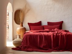 a bed with red sheets and pillows in a white room next to a large vase