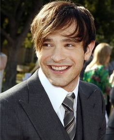 a man in a suit and tie smiling at the camera while people are taking pictures behind him