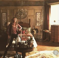 a woman standing next to a bed in a room with wooden paneling and chandelier