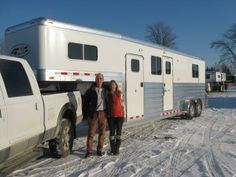 two people standing in front of a horse trailer on snow covered ground with trees behind them