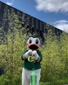 the mascot is standing in front of some tall grass and bushes, with a green bottle next to it